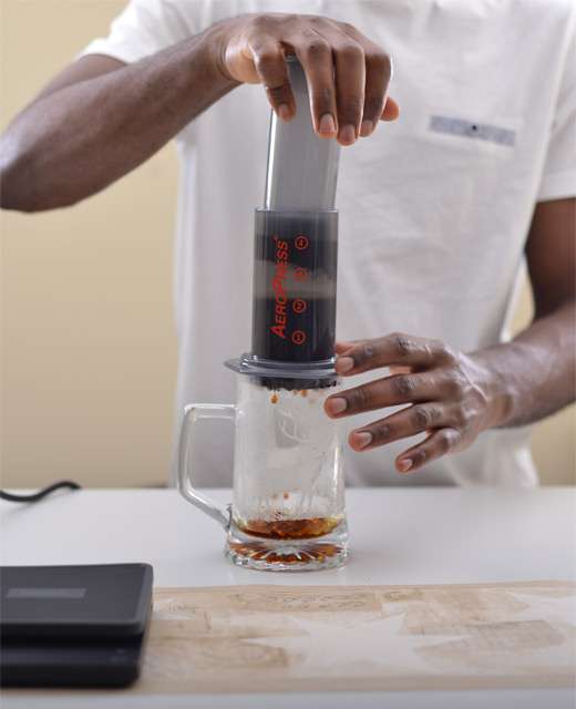The hands of a barista are seen applying steady pressure on the AeroPress plunger, extracting the coffee smoothly into the awaiting mug below.