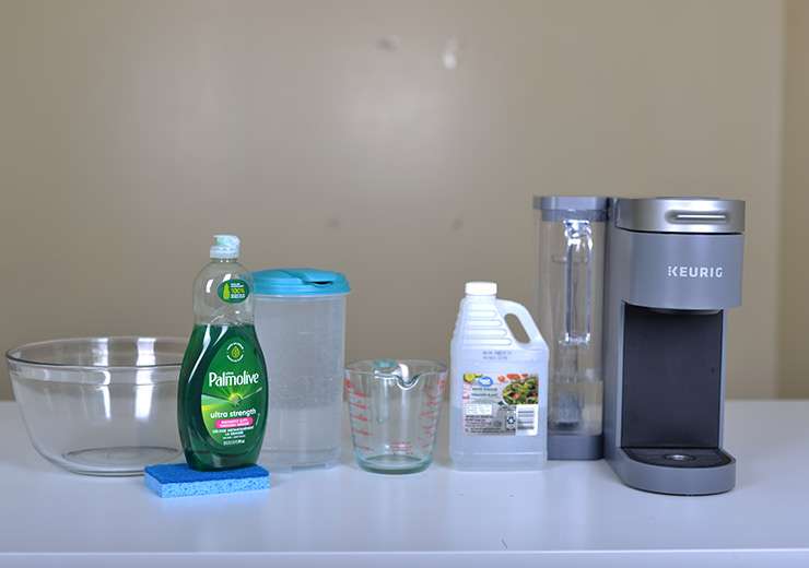 Keurig K-Supreme coffee maker descaling materials on a table: Keurig machine, vinegar, water, ceramic mug, measuring cup, dish soap, and sponge